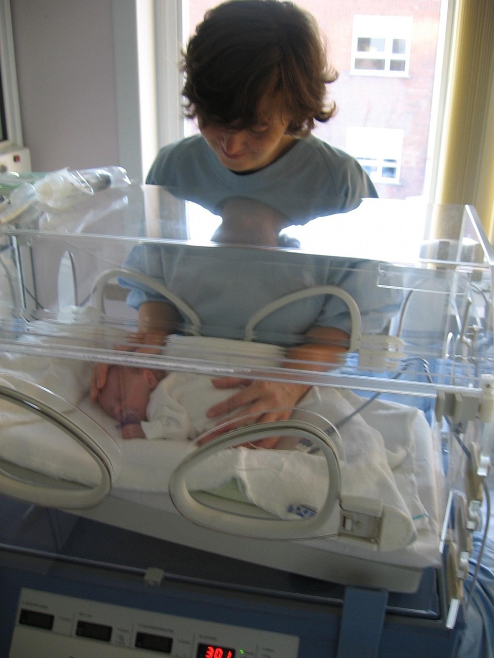Mama in hospital smock places hands on baby in incubator