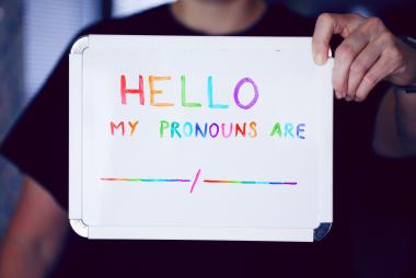 Person in black T-shirt holds whiteboard with multicoloured lettering: "Hello my pronouns are"